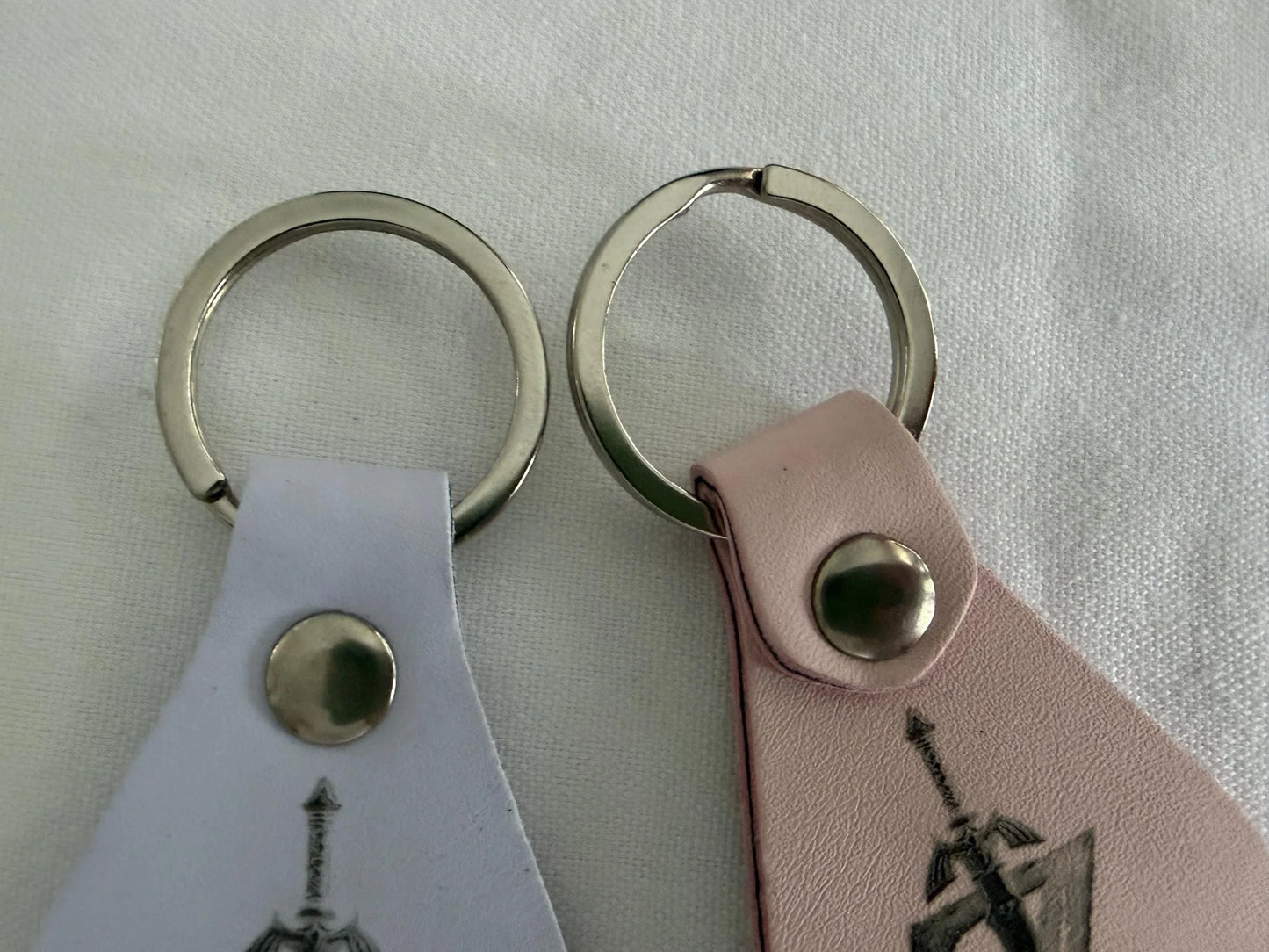Two leather keychains with metal rings, one white and one pink, on a fabric surface