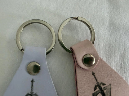 Two leather keychains with metal rings, one white and one pink, on a fabric surface