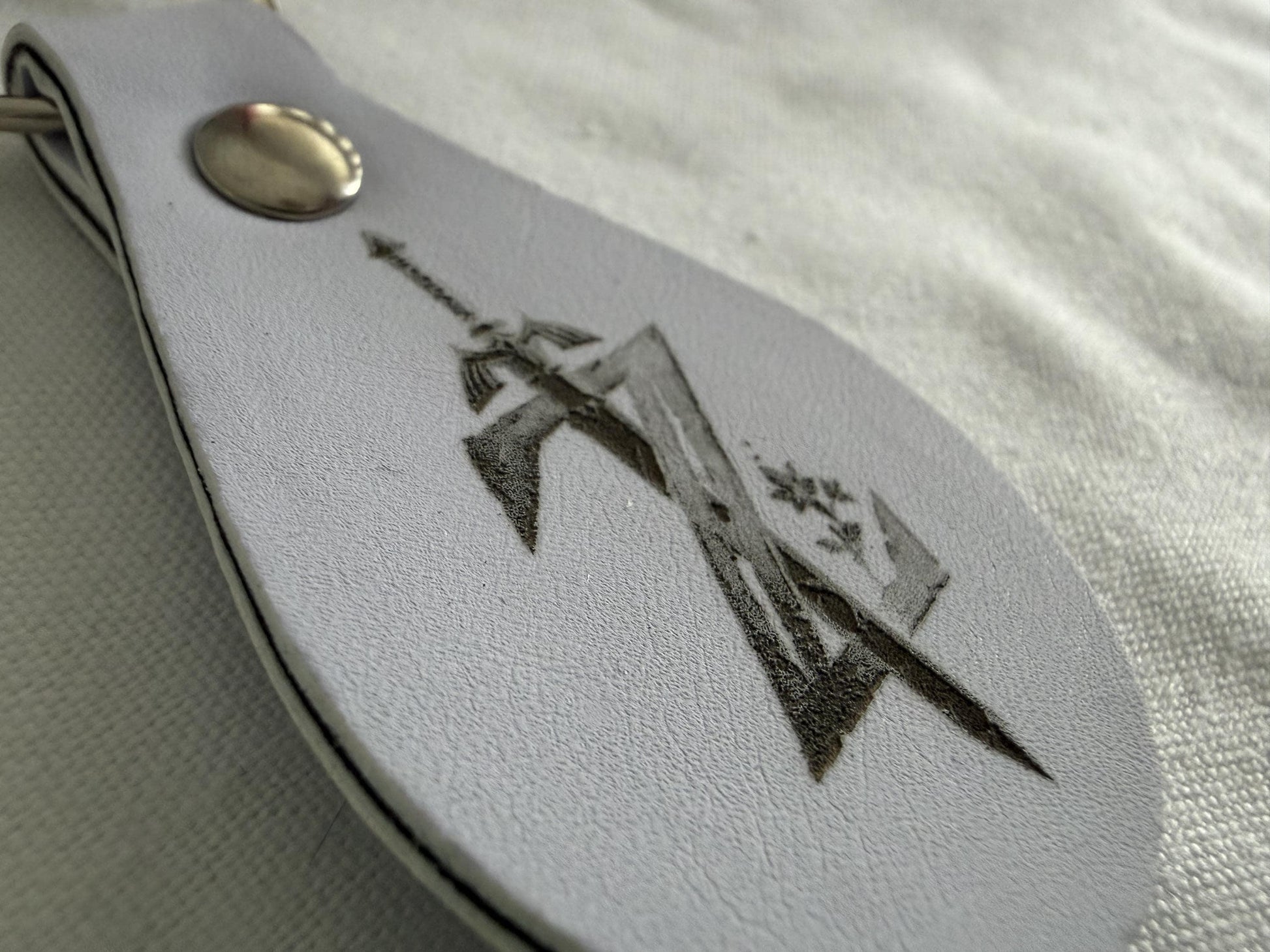 White leather keychain with engraved sword and shield, metal snap closure on fabric surface