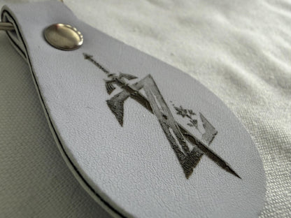 White leather keychain with engraved sword and shield, metal snap closure on fabric surface