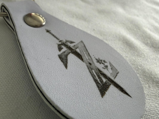 White leather keychain with engraved sword and shield, metal snap closure on fabric surface