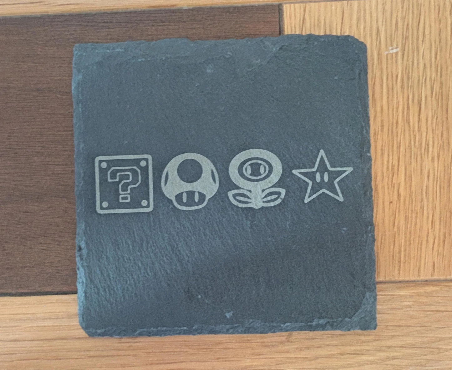 Square engraved slate coaster with four retro game icons on a wooden surface